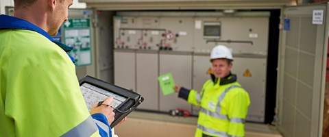 Zwei Techniker in Warnschutzjacken mit portablem Eingabegerät vor offener Trafostation Zwei Techniker in Warnschutzjacken mit portablem Eingabegerät vor offener Trafostation