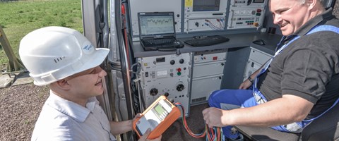 Lachender Arbeiter mit Helm und portablem Gerät vor einem offenen Technikfahrzeug, in dem ein Kollege sitzt Lachender Arbeiter mit Helm und portablem Gerät vor einem offenen Technikfahrzeug, in dem ein Kollege sitzt