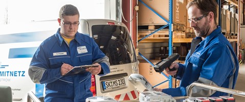 Zwei Arbeiter, beide im Blaumann, erfassen in einem Lager mit Scanner und Klemmbrett technische Geräte auf einem Tisch Zwei Arbeiter, beide im Blaumann, erfassen in einem Lager mit Scanner und Klemmbrett technische Geräte auf einem Tisch