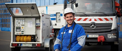 Mitarbeiter trägt Arbeitskleidung mit Helm und ein Bedienmodul um den Hals - er steht vor einem Lkw mit Kran und einem offenen Anhänger mit technischem Gerät Mitarbeiter trägt Arbeitskleidung mit Helm und ein Bedienmodul um den Hals - er steht vor einem Lkw mit Kran und einem offenen Anhänger mit technischem Gerät