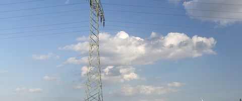 Hochspannungsmast auf einer grünen Wiese mit Windrädern unter blauem Himmel mit weißen Wolken Hochspannungsmast auf einer grünen Wiese mit Windrädern unter blauem Himmel mit weißen Wolken