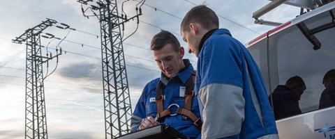 Zwei Monteure in Blaumännern blicken, am Heck eines Fahrzeugs vor zwei Hochspannungsmasten stehend, auf ein portables Eingabegerät Zwei Monteure in Blaumännern blicken, am Heck eines Fahrzeugs vor zwei Hochspannungsmasten stehend, auf ein portables Eingabegerät