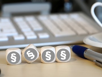 Vier Holzwürfel auf einem Schreibtisch neben einem Kugelschreiber vor einer Tastatur zeigen alle das Paragrafenzeichen Vier Holzwürfel auf einem Schreibtisch neben einem Kugelschreiber vor einer Tastatur zeigen alle das Paragrafenzeichen